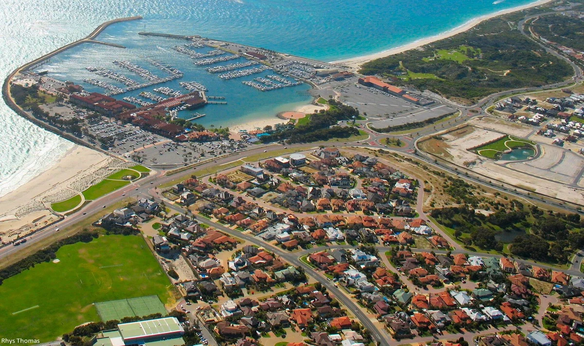 Aerial view of Hillarys Boat Harbour, its curved breakwater packed with moored boats, the sheltered swimming beach inside it, and the open surf beach and suburban streets of Sorrento stretching away to the south