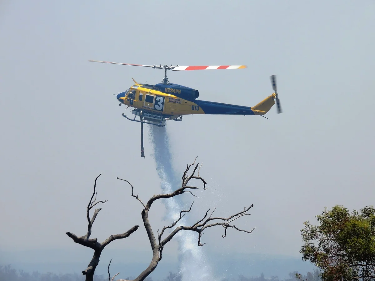 A yellow water-bombing helicopter drops water over smoky bushland