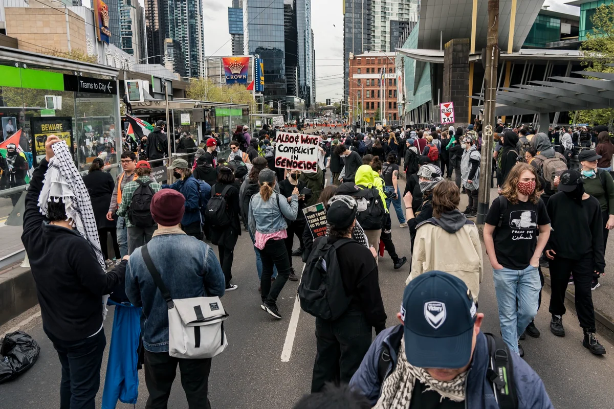 A crowd of protesters, some in keffiyehs, fills a Melbourne street outside the convention centre; one holds a sign reading Albo and Wong complicit in genocide