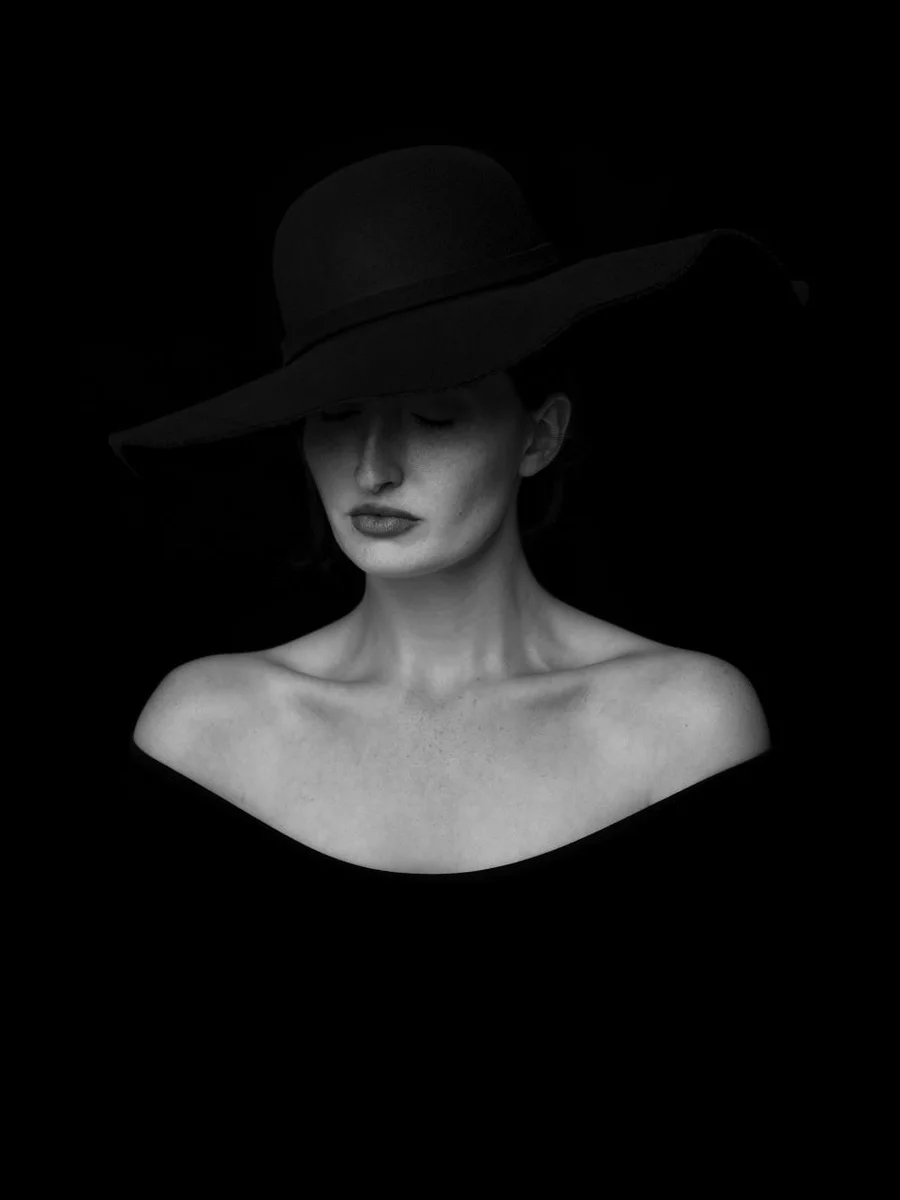 A woman in a wide-brimmed black hat photographed in black and white against a black background